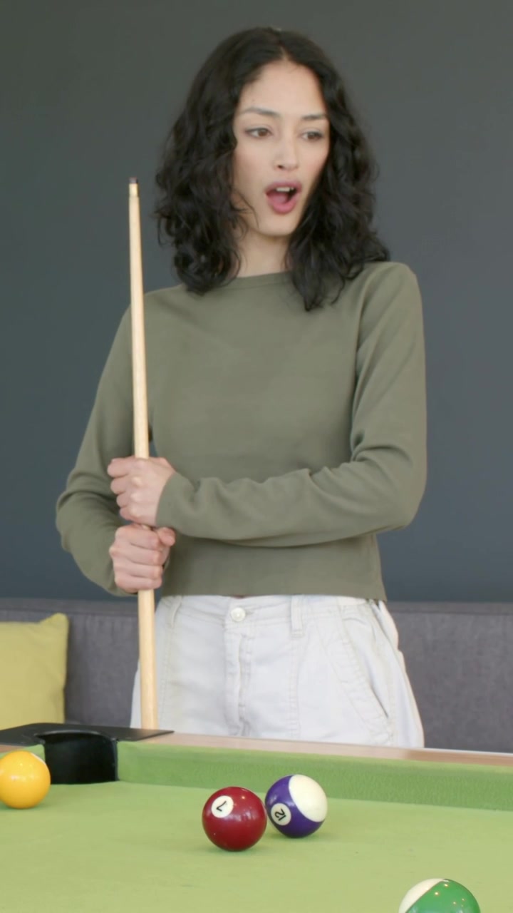 Vertical video: Holding woman in olive top noticing ball cluster near pocket, gripping cue at home