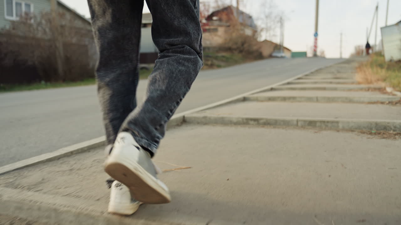 Scarpe da ginnastica che scendono dal marciapiede, camminando lungo una strada suburbana verso un'officina di riparazione, cappotto color cammello e zaino, passo attento sul bordo rialzato del marciapiede, pavimentazione usurata, foglie sparse, case distanti, luce autunnale.