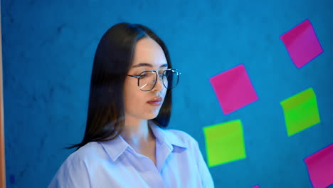 dark-haired woman wearing glasses and a shirt puts stickers on the glass board. business concept.