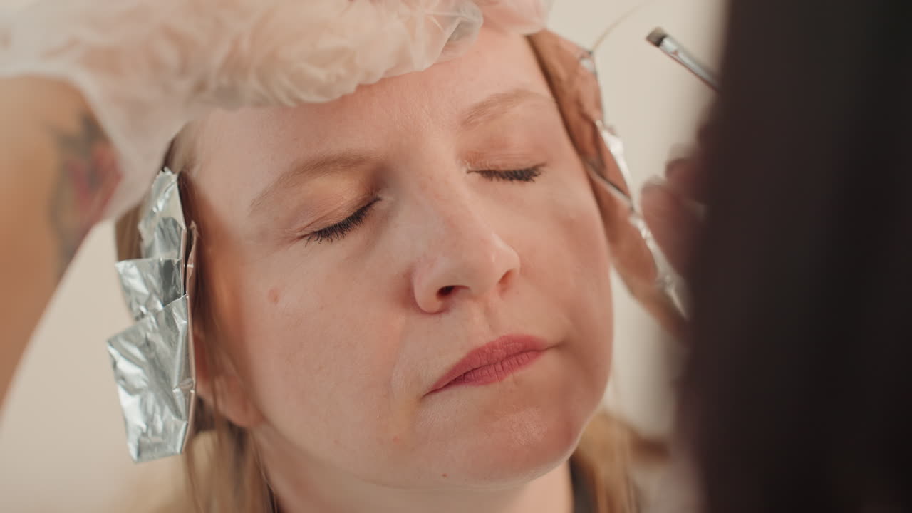 Kvinde modtager microblading-behandling af øjenbryn, lukkede øjne, tekniker med handsker bruger microblade til at kortlægge og deponere pigment langs bryn, staniol i håret, neutral studiebelysning, roligt afslappet udtryk