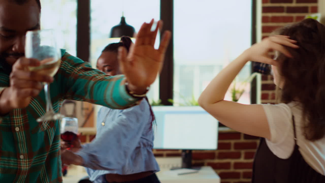 Joyful friends dancing at house party while drinking wine glasses, enjoying celebration