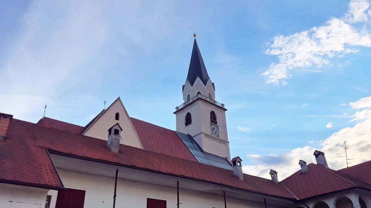 Chiesa di San Canziano nel centro storico di Kranj, Slovenia, con tetti di tegole rosse, torre dell'orologio, guglia gotica e un cielo serale di un blu brillante.
