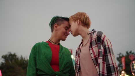 a blonde girl with short hair in a plaid shirt and a girl with green hair in a green shirt keep their faces close and look at each other during a date in an amusement park