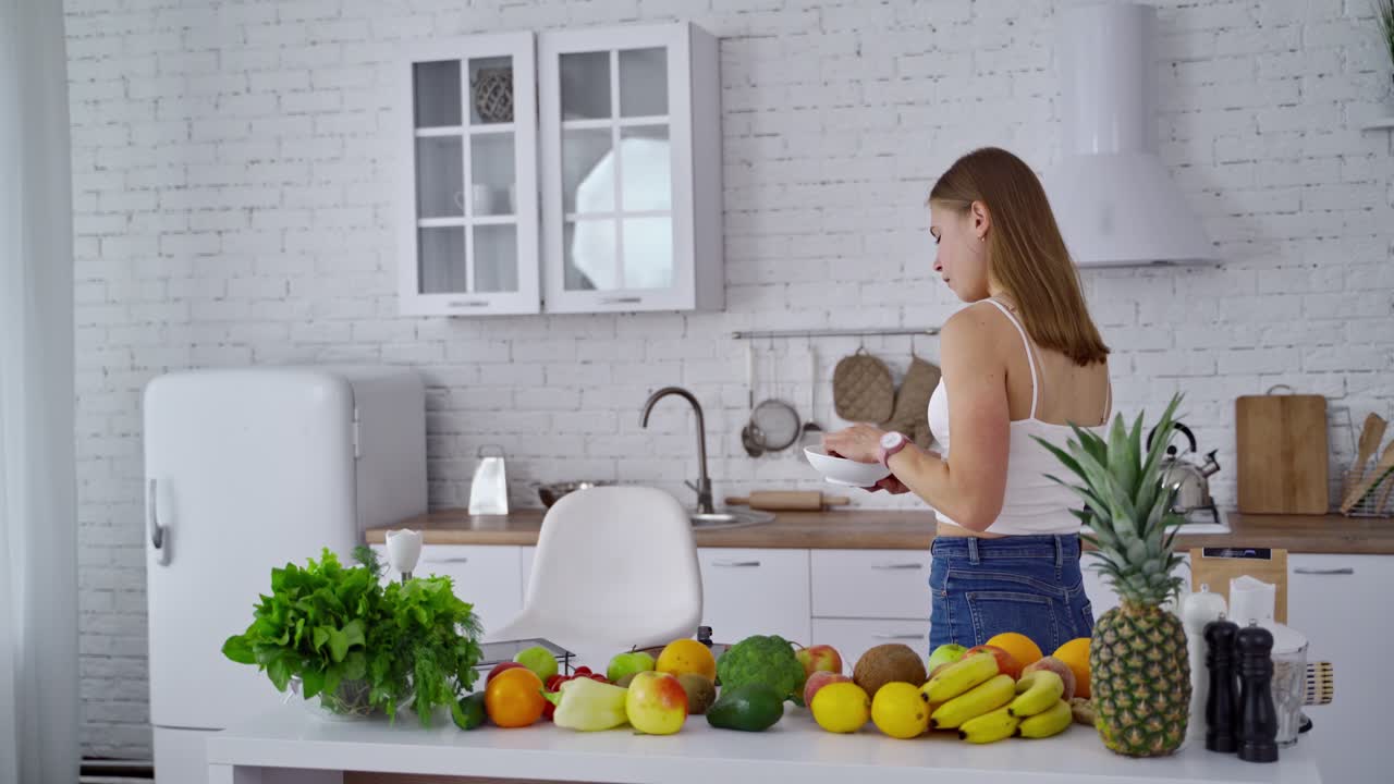 Side view of a pretty woman in the kitchen. Kitchen table full of fresh organic fruits and vegetables for healthy diet. Woman eating dietetic food at home.