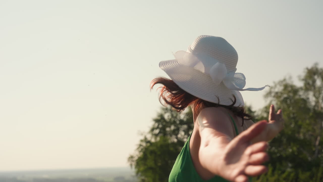 woman wearing white sunhat in green dress dances gently on scenic hilltop with calm river and distant forest in background under warm sunlight