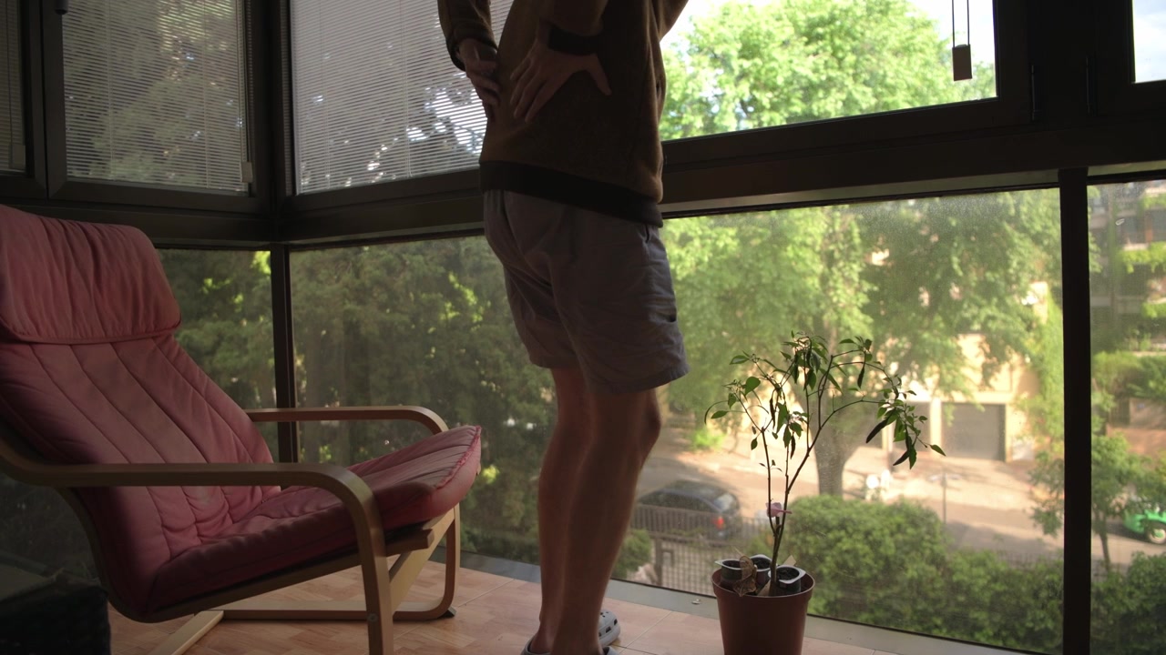 Un uomo siede su un balcone interno, godendosi la vista sul giardino. Si alza e si stira, ammirando il verde esterno. Ideale per temi di lifestyle o relax