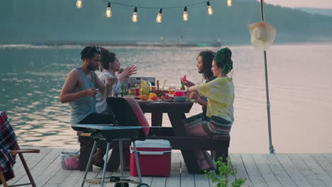 friends eating and drinking at dinner party on lake pier