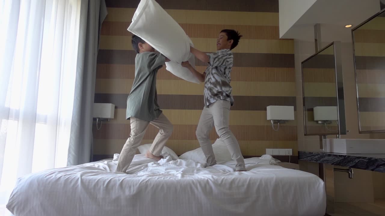 Two brothers are enjoying pillow fight on bed in apartment laughing having fun together. Leisure time, happy family and joyful people concept.