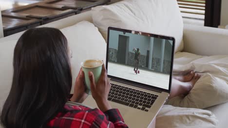 Composite-of-woman-sitting-at-home-holding-coffee-watching-athletics-running-event-on-laptop