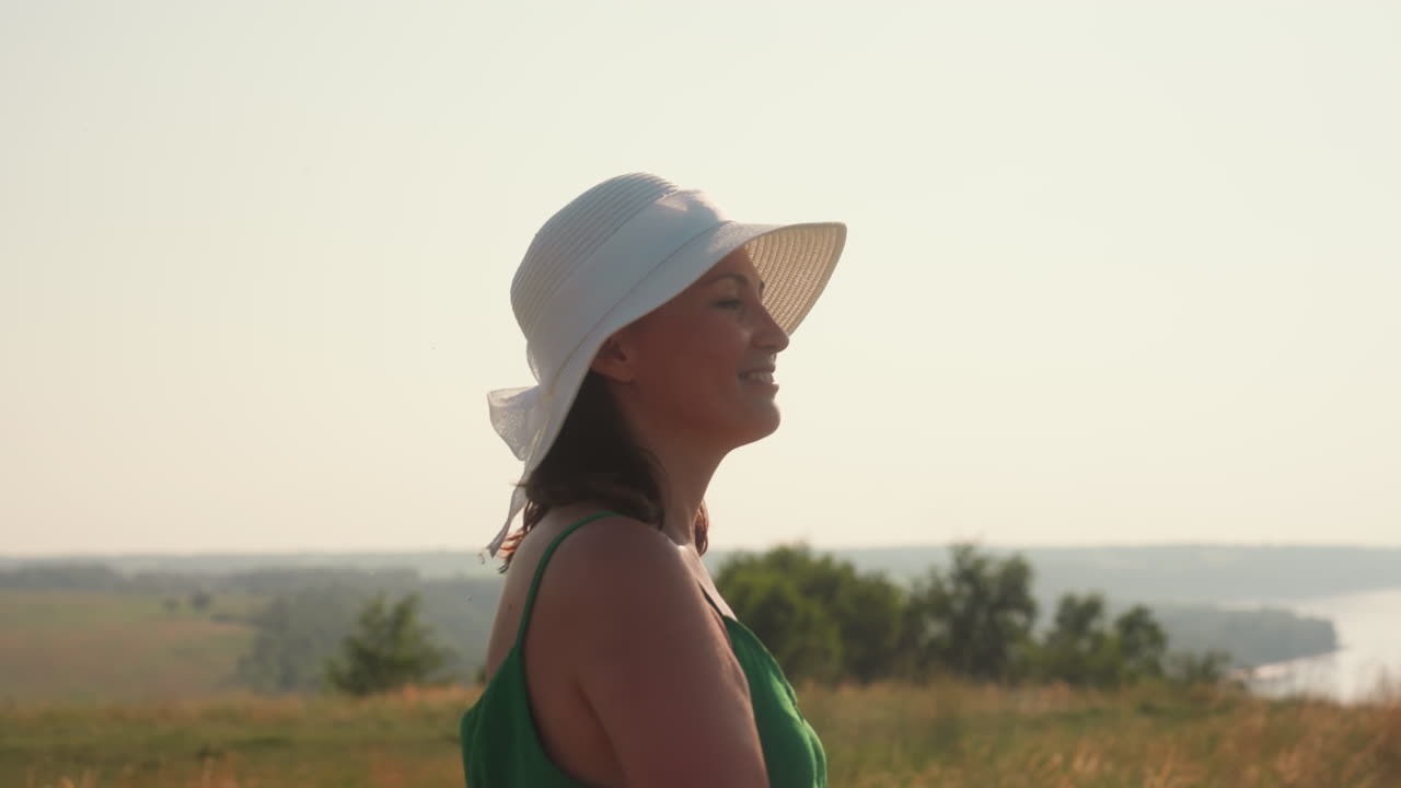 Jovem mulher com chapéu de sol branco e vestido verde gira alegremente com os olhos fechados sob o sol brilhante em uma colina gramada aberta com árvores suaves e uma vista tranquila do rio ao longe.