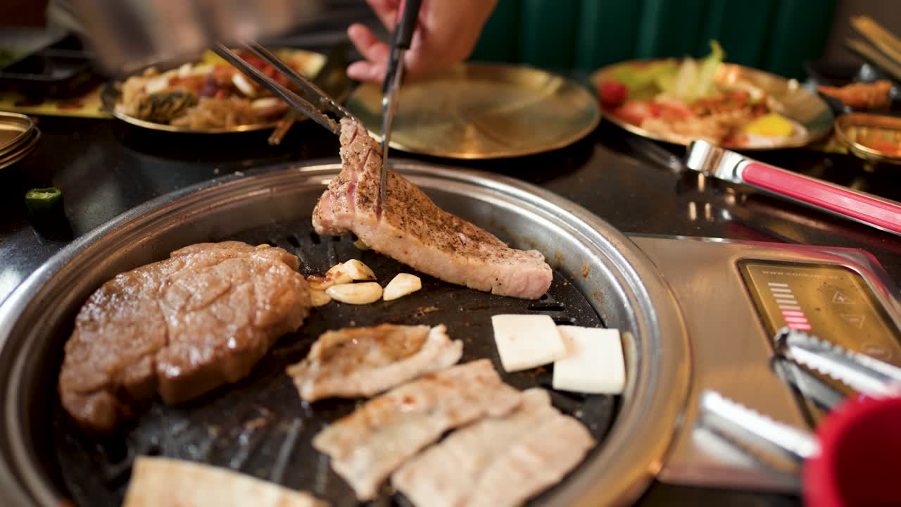 Hand uses tongs and scissors to cut pork belly on tabletop grill, warm indoor lighting