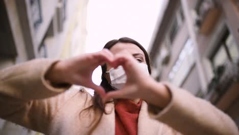 beautiful girl wearing protective medical mask and fashionable clothes does heart shape at street