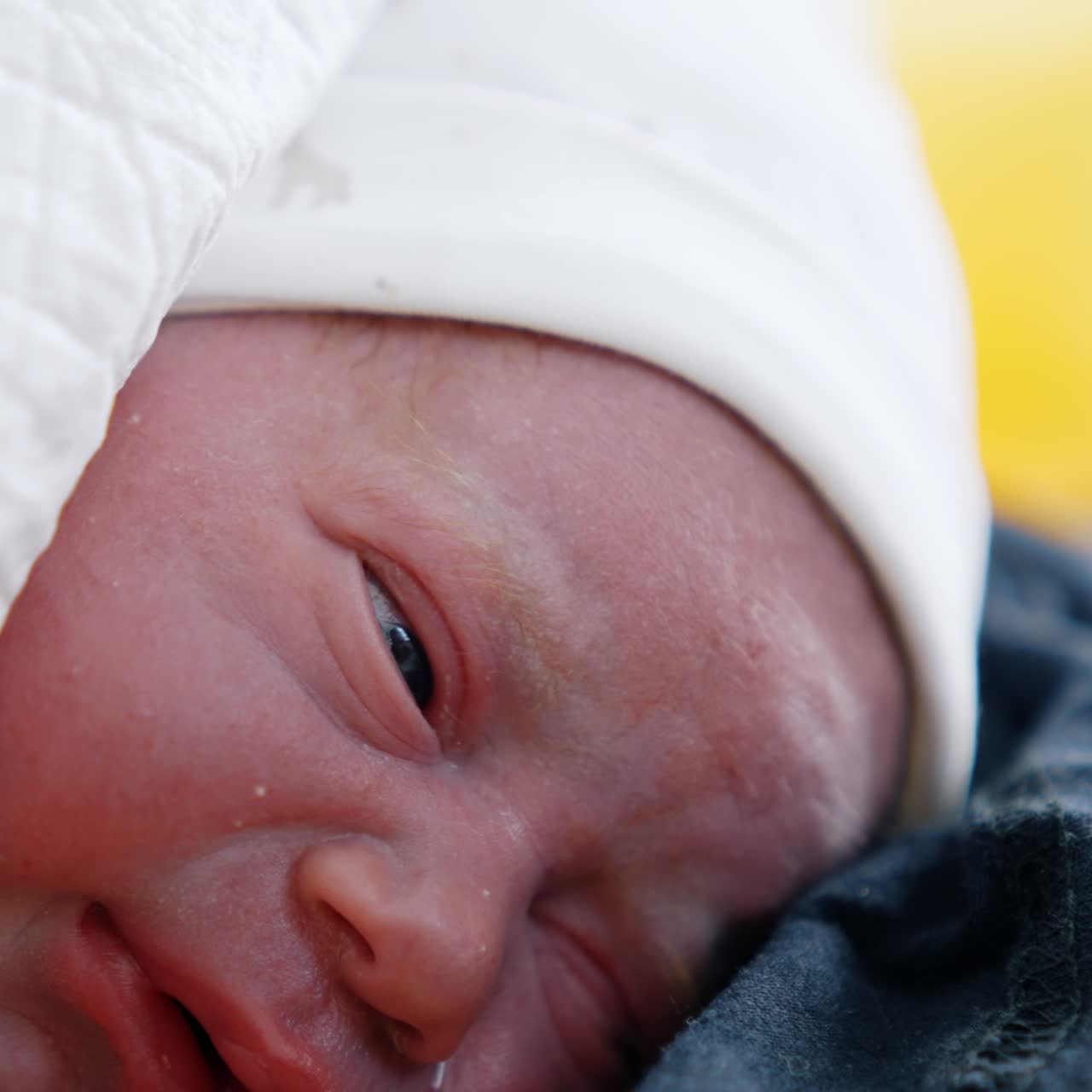 Face of a newborn right after birth. Tiny baby in white cap wants to cry and blinking eyes. Close up