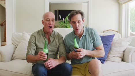 Portrait-of-happy-caucasian-grandfather-with-adult-son-with-beer-watching-tv-in-living-room