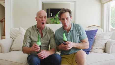 portrait of happy caucasian grandfather with adult son with beer watching tv in living room