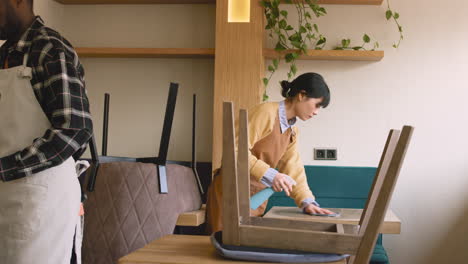 waitresss cleaning coffee shop table with disinfectant spray and rag while waiter arranging chairs