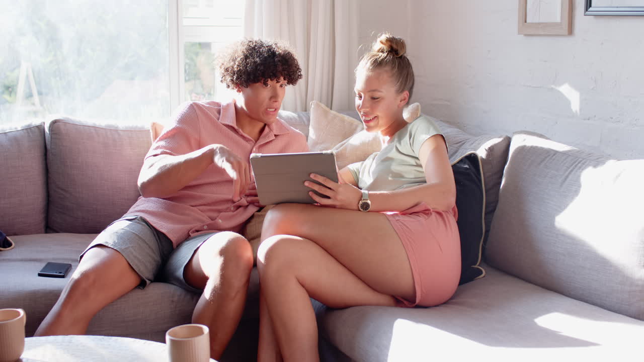 Smiling diverse couple relaxing on sofa, enjoying tablet together in bright living room, at home