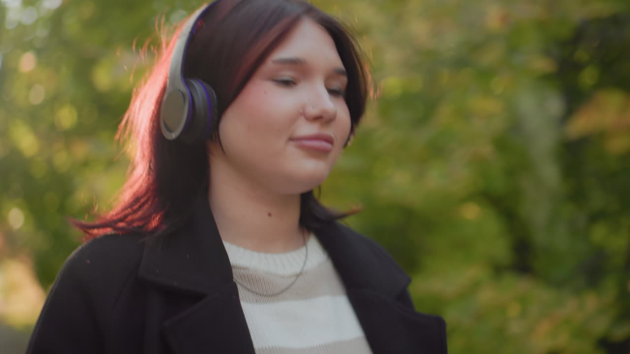 Medium view of carefree girl wearing headphones, holding yellow leaves, gazing around while strolling through sunlit autumn park, relaxed mood, soft breeze, warm light, gentle smile, nature ambience