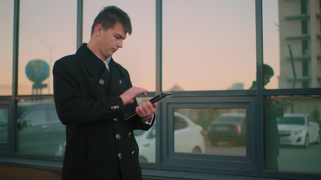 ceo는 유리 사무실 건물 근처에서 걸어가는 동안 태블릿을 신중하게 작동합니다. 유리 위에 반사되는 것은 흐릿한 건물, 주차된 자동차 및 세계 지도 구조를 보여줍니다.