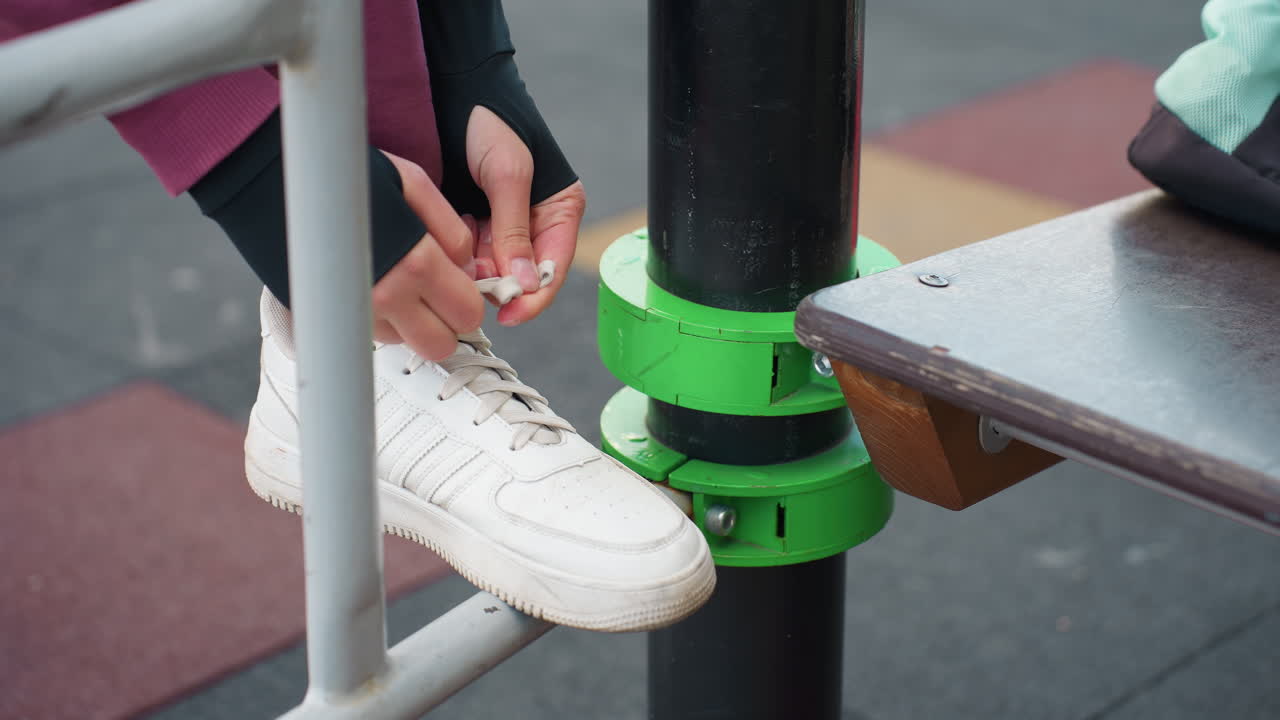 Female feet perched on iron exercise bar tying canvas sneaker laces with careful focus before dynamic calisthenics routine under open sky in urban fitness park surrounded by modern apartment view