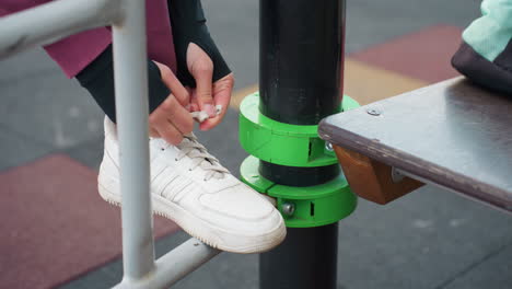 female feet perched on iron exercise bar tying canvas sneaker laces with careful focus before dynamic calisthenics routine under open sky in urban fitness park surrounded by modern apartment view