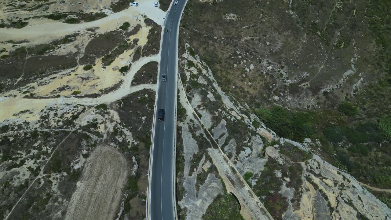 Aerial tilt down follows cars driving winding Gozo road between fields and hills with coastline visible in the distance, Malta