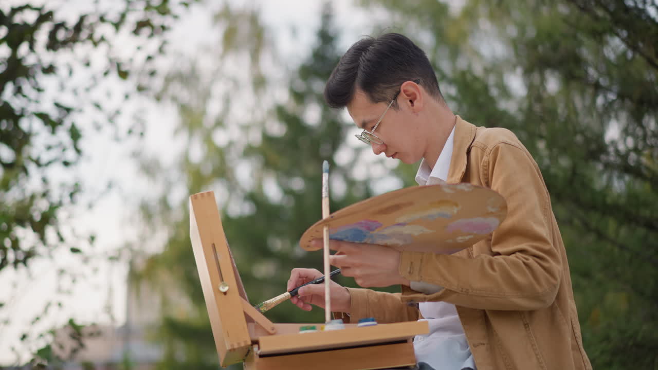 Artista asiatico che dipinge all'aperto al cavalletto, abbozzando un paesaggio verdeggiante del parco e mescolando colori a olio su una palette di legno mentre studia la composizione. Uomo concentrato con giacca marrone chiaro che aggiusta le pennellate, cattura