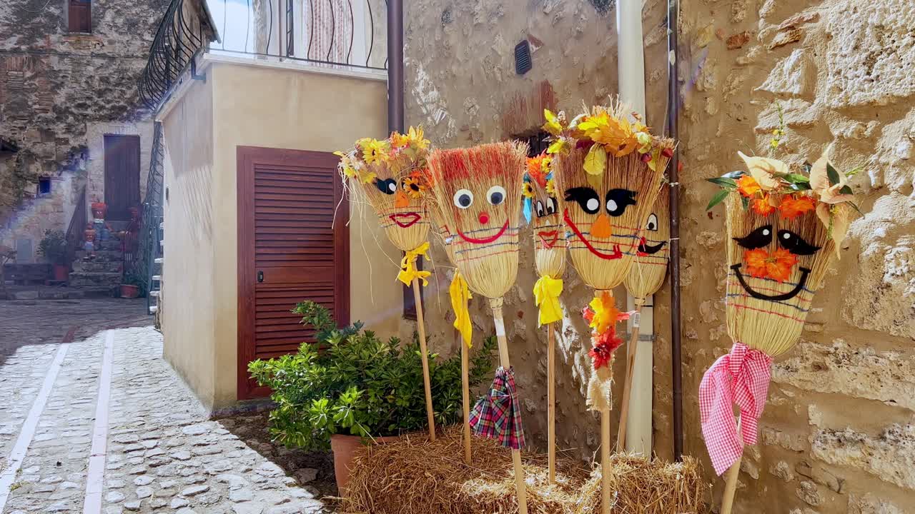 Brooms decorated with whimsical, happy faces and flowers line a narrow cobblestone street during a traditional harvest festival in Italy