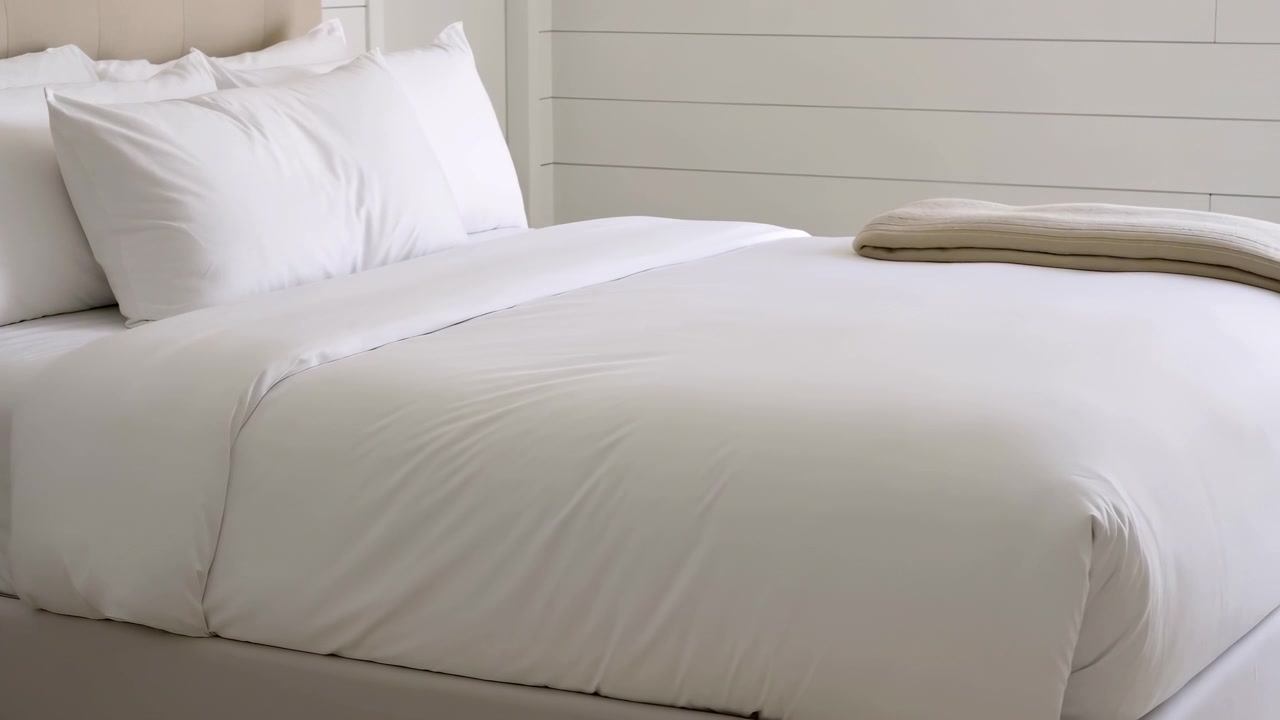 Low-angle bedroom view showing neatly made bed with taut white sheet, smooth duvet folded back slightly, four fluffed pillows, upholstered beige headboard, and folded tan blanket in soft daylight