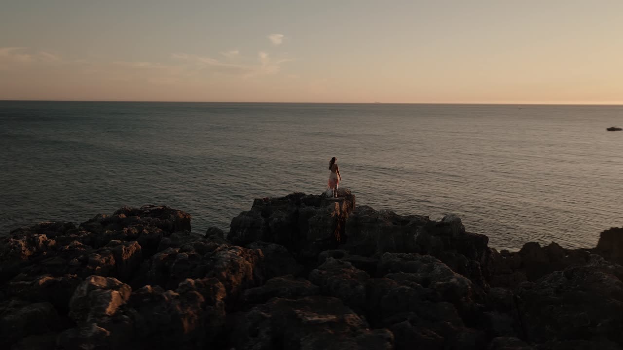 Orbita aerea di una donna con un abito svolazzante ferma su scogliere rocciose, di fronte al vasto mare durante l'ora d'oro in Portogallo.