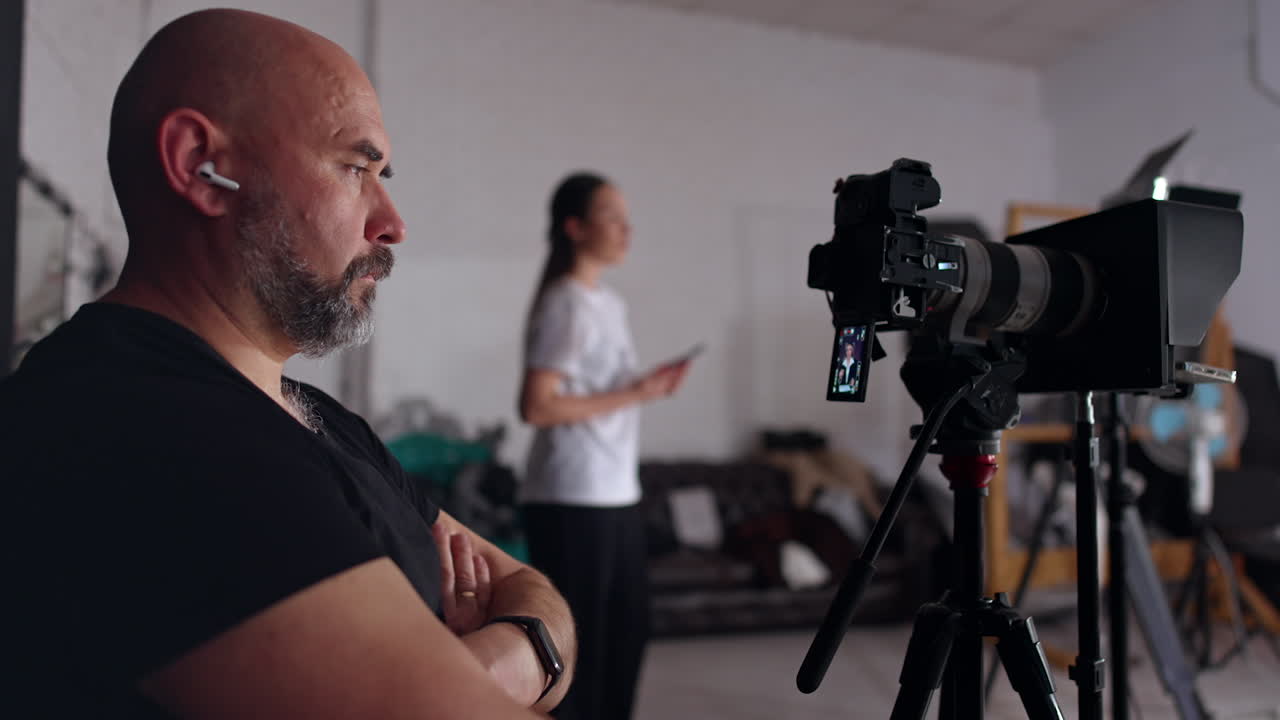 Serious man in black t-shirt sits his hands folded on chest in front of camera. Cameraman looks focused on the camera display. Female stands at blurred backdrop.