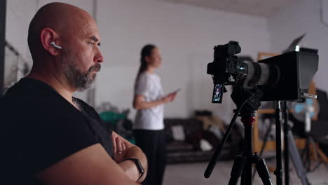 serious man in black t-shirt sits his hands folded on chest in front of camera. cameraman looks focused on the camera display. female stands at blurred backdrop.