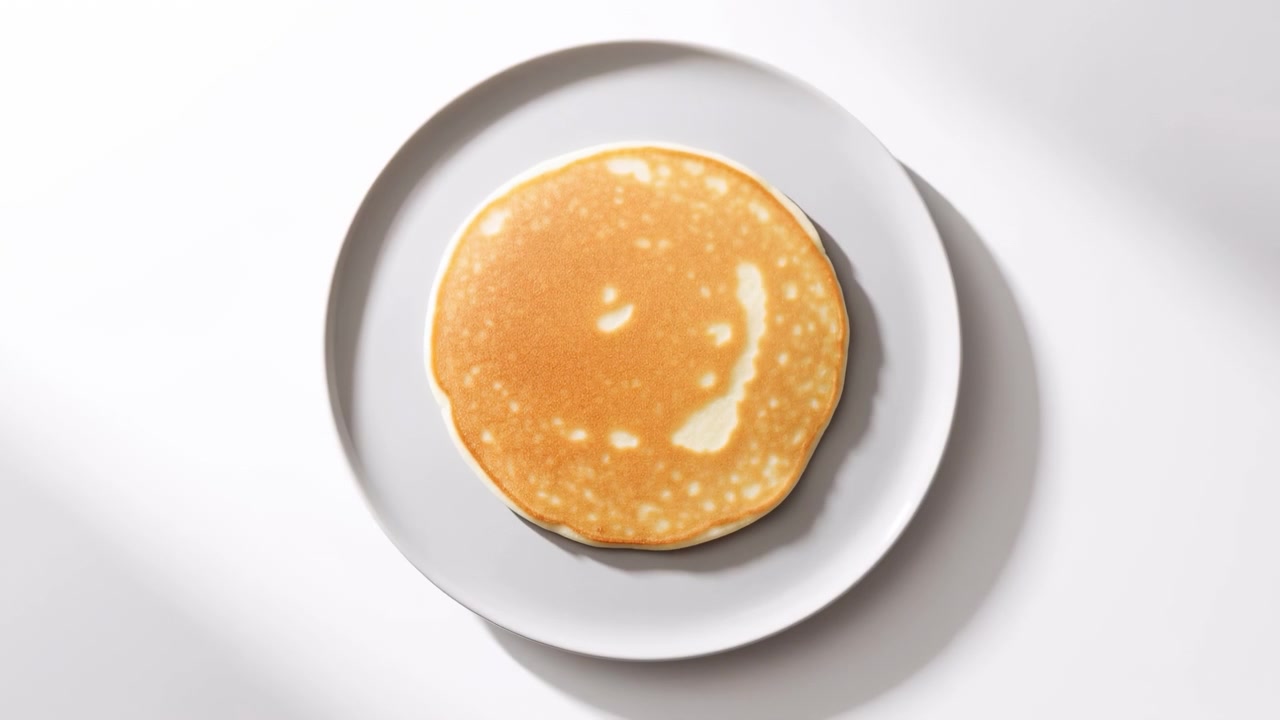 Golden pancake rises, turns in air, then settles flat on white porcelain plate in bright overhead breakfast scene with soft light, minimal styling, and clean tabletop, with pancake, plate, white