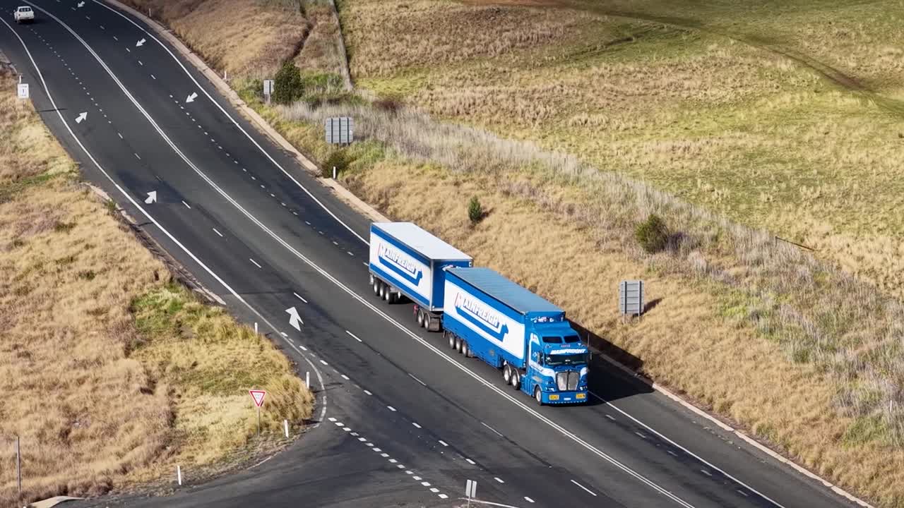 Un semirimorchio blu con rimorchi doppi percorre un'autostrada rurale attraverso una campagna arida