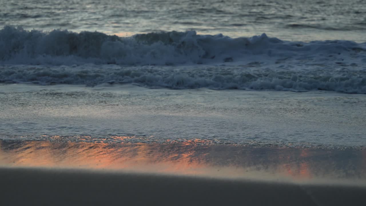 Close up footage of the sea rolling in, gently lit up by a warm orange sunset, captured in 60fps, slow motion
