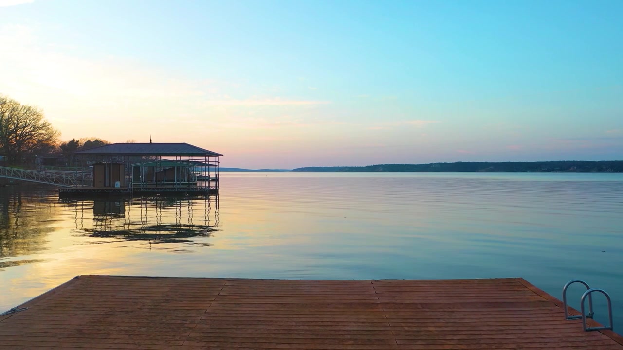 Droneoptagelse over Branson, Missouri ved solnedgang, med roligt Ozark-søvand, skovklædt kystlinje, huse ved vandet, overdækkede bådbroer, pastelfarvet himmel og gyldne refleksioner over reservoiret.