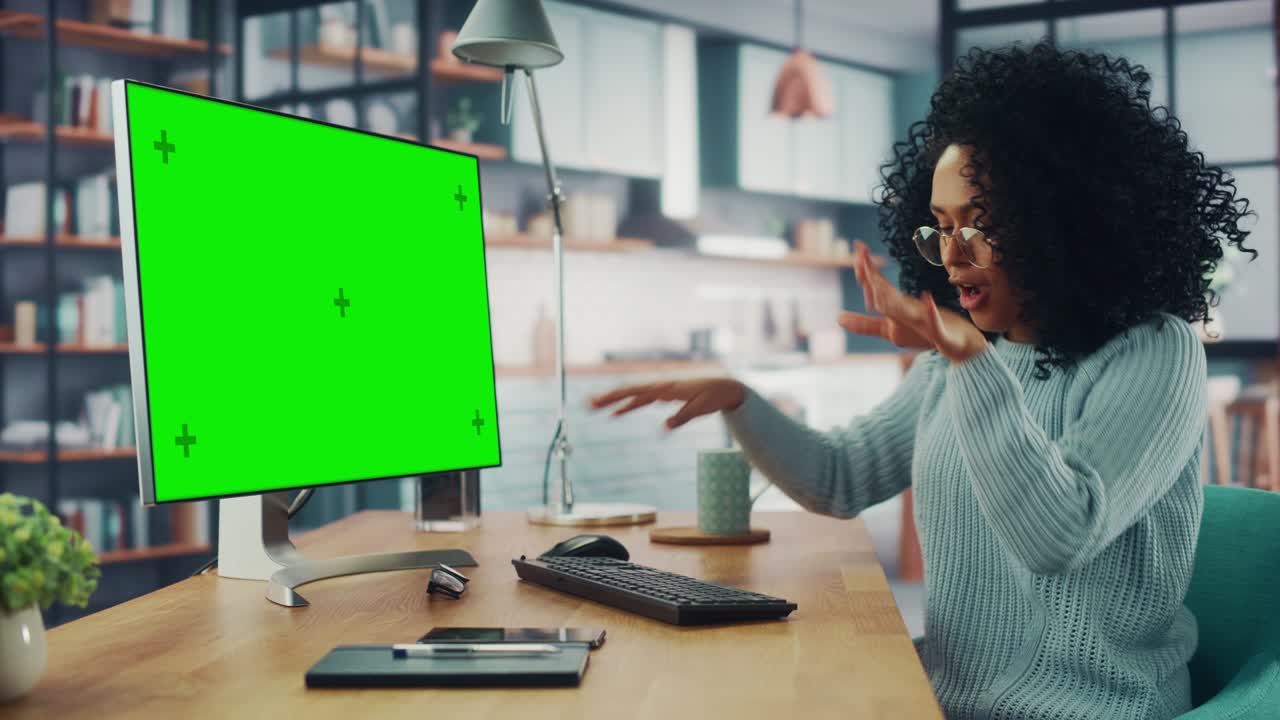 Latina Female Specialist Dancing While Sitting Next to Desktop Computer with Green Screen Mock Up Display at Home Living Room. Freelancer Female Having Fun while Remotely Working on a Project.
