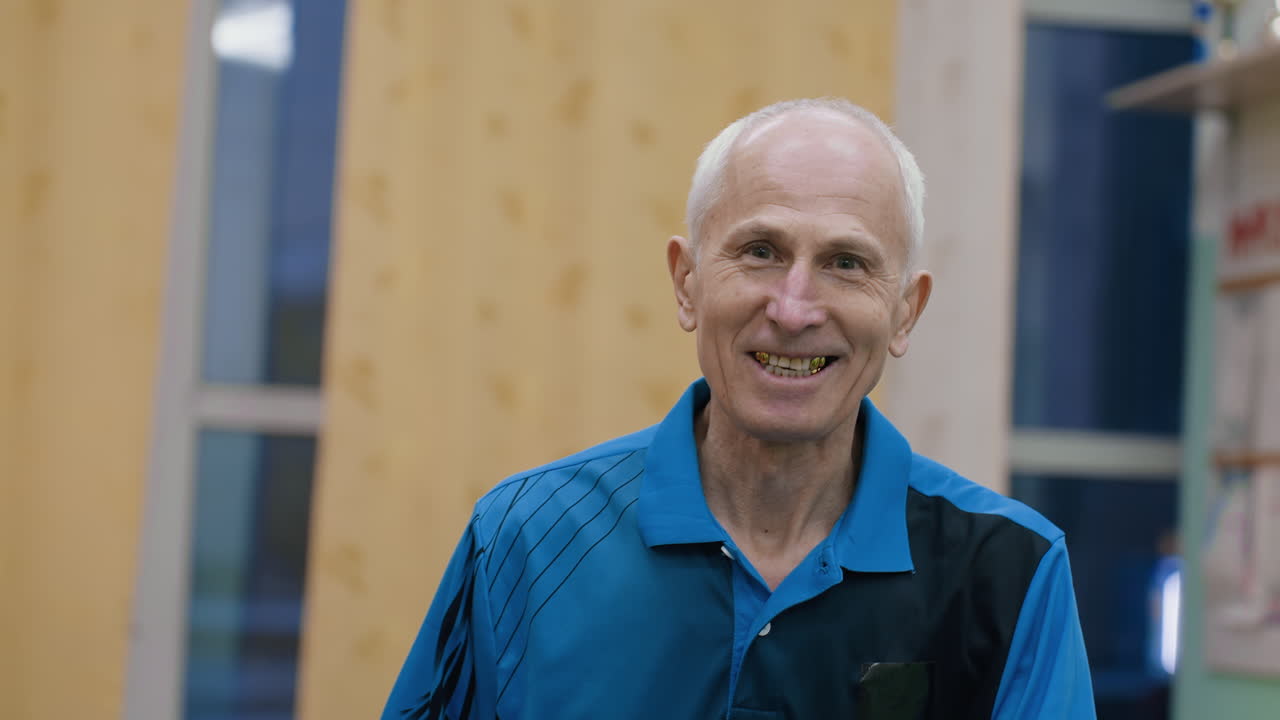 Aged man in blue sportswear attempts playful trick by holding table tennis ball with air breath, laughing joyfully as ball hovers briefly before falling during indoor match atmosphere