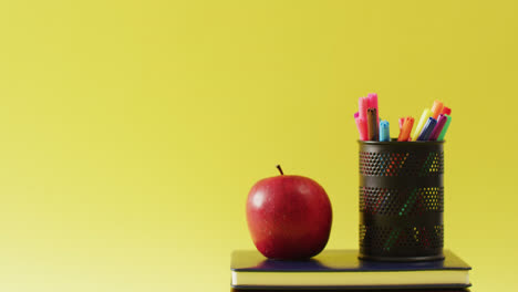 video of crayons in container, books and apple on yellow surface