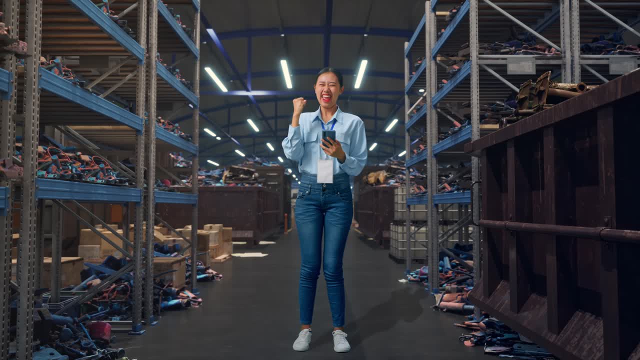 Mulher asiática de corpo inteiro com seu smartphone, em um armazém industrial de sucata e reciclagem, levanta o punho com um grito de vitória.