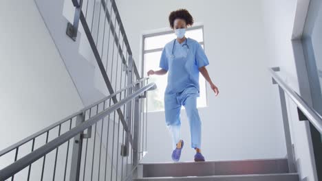 Mixed-race-female-doctor-wearing-face-mask-running-down-stairs-in-hospital