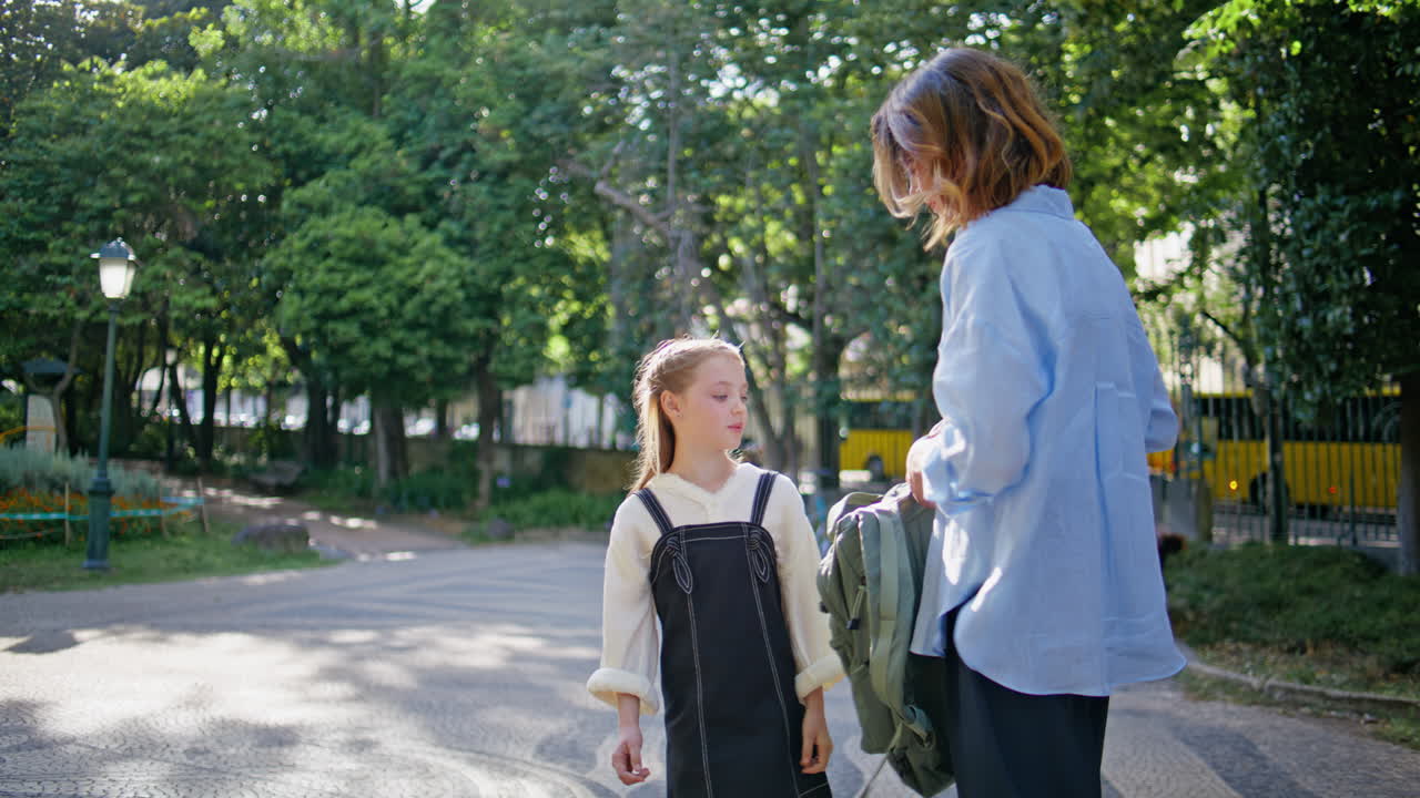 Mother daughter talking greenery square. Caring woman taking backpack at park