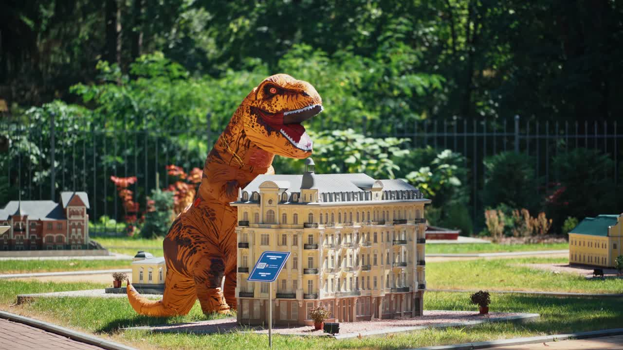 Big dragon costume in artificial city. Person in funny blow-up suit standing close to handmade multi-storey building in the park.