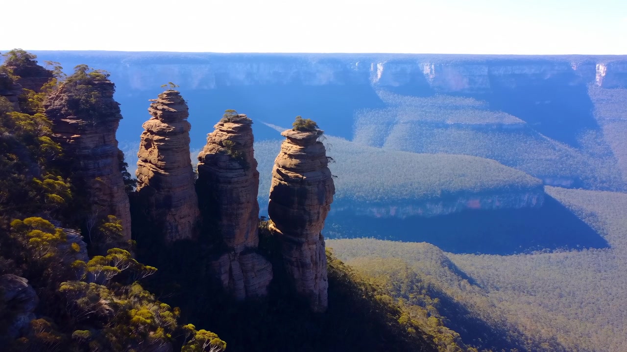 Quatro altos pilares de arenito erguem-se de um cume de falésia vegetado ao lado de um amplo vale azul, com escarpa de topo plano, horizonte nebuloso, estratos quentes e forte contraste em uma paisagem remota.