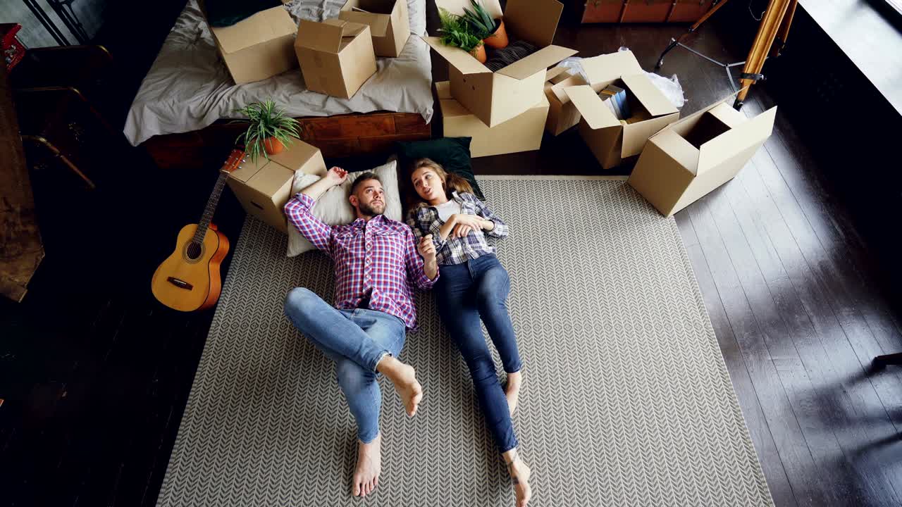 vista superior de una pareja amorosa acostada en el suelo del dormitorio en un nuevo apartamento y hablando. guitarra, cosas empacadas en cajas de cartón, cama doble y alfombra moderna son visibles.