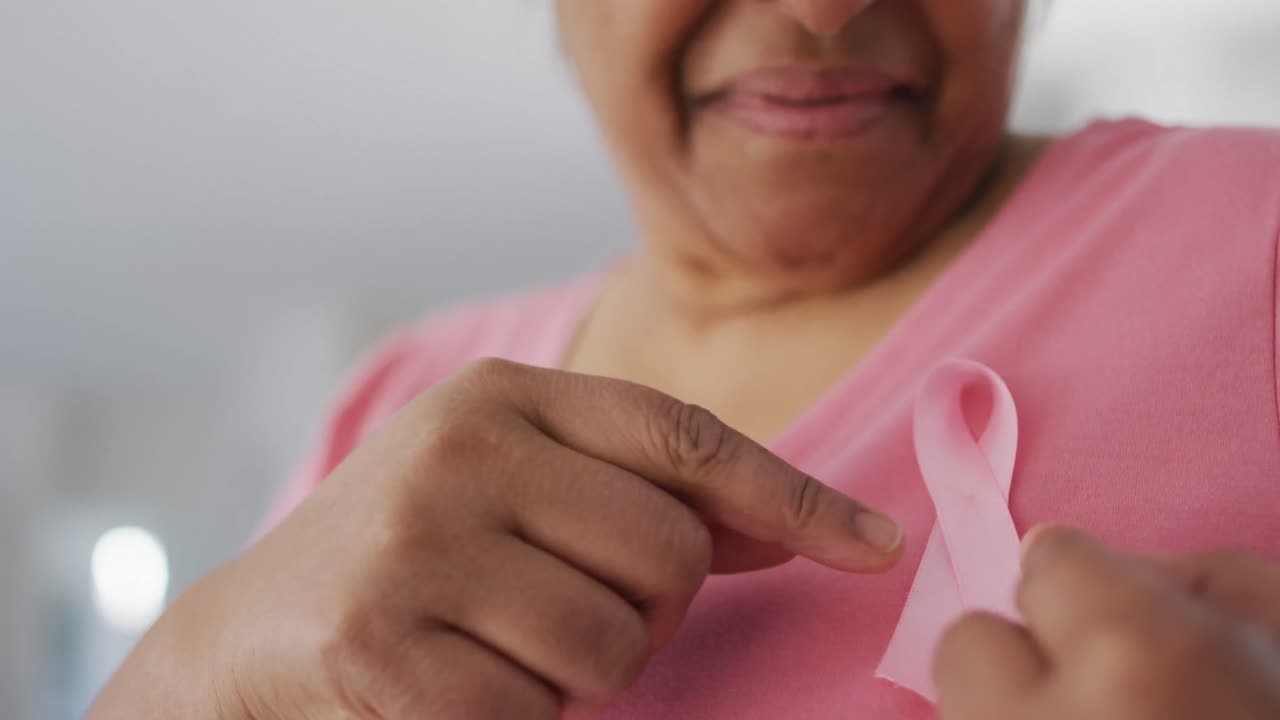 Meio-corpo de mulher idosa afro-americana sorrindo, com camiseta rosa e fita rosa de conscientização sobre o câncer
