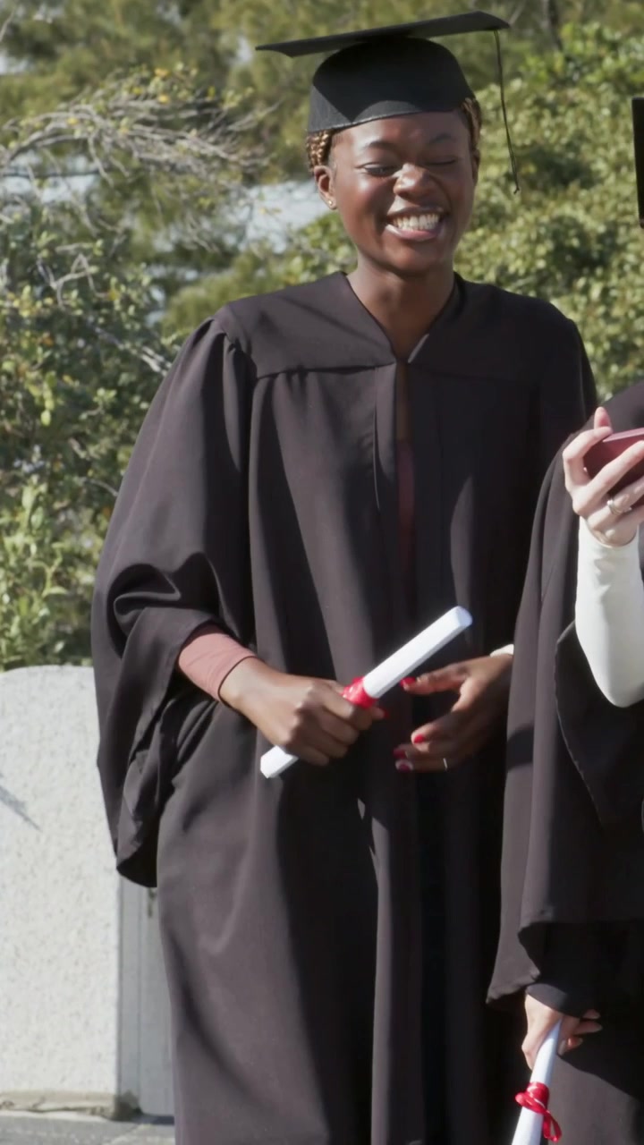 Vertical video: Raising pink phone grad in black gown cap taking selfie on campus rail with diploma