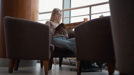 young woman reading book while seated at a cozy cafe table with coffee cup, she appears absorbed in her reading with a calm and focused expression, soft natural light through large windows