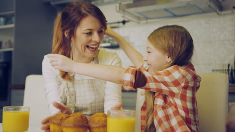 good looking mother in the apron coming to her daughter with muffins on the plate and thay both smelling them in the kitchen at the table. inside