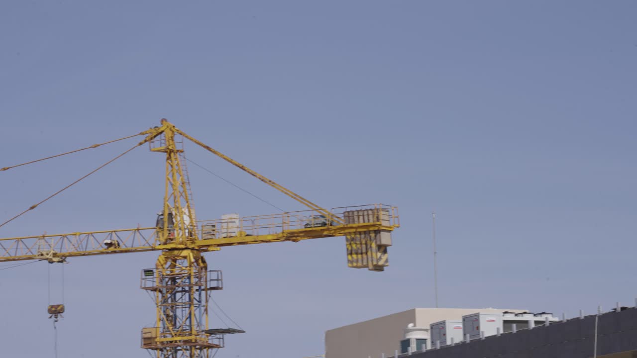 A large, yellow tower crane stands tall against a clear blue sky at a busy construction site. The crane is essential for building a new high-rise structure